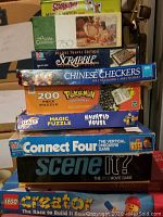 Stack of assorted board games and puzzles including Scooby-Doo Match, 750-piece puzzle, Scrabble Deluxe Travel Edition, Chinese Checkers, 200-piece Pokemon puzzle, Galt Magic Puzzle Haunted House, Connect Four, Scene It?, and LEGO Creator.