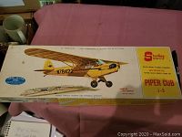 Top view of the Sterling Piper J3 Cub model kit box showing illustration of yellow plane, model info sticker, and brand logo