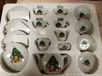 17-piece vintage porcelain Christmas tea set packed in a foam tray showing cups, saucers, sugar bowl, creamer, and small serving bowls decorated with Christmas trees, teddy bears, and gifts.