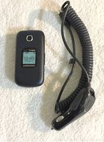 Samsung flip phone closed showing external screen with time and Verizon branding next to coiled cord car charger.