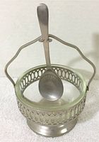Front view of the vintage jam condiment server showing metal frame with cut-out design, clear glass insert bowl, and handle with spoon placed inside.