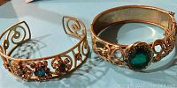Photo showing both gold-tone costume jewelry bracelets, one open cuff style with multiple amber-colored stones and one hinged bangle with a large green stone and scroll designs.