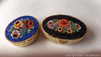 Two oval Italian micro mosaic pill boxes with floral designs, one with blue background and one with black background on gold-tone metal bodies
