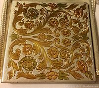 Close-up of the compact top showing gold and bronze scrollwork and flowers on textured silver background.