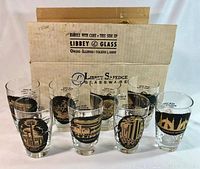 Set of eight clear tapered iced tea glasses with gold and black World's Fair designs arranged in front of the original cardboard box labeled Libbey Safedge Glassware Owens Illinois Toledo Ohio.
