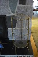 Full view of the brass and glass three-tier side table against a stone wall and black hearth tile floor, showing round glass shelves and brass frame.