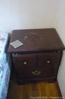 Full view of wooden end table positioned next to a bed with visible surface discoloration and a coaster on top.