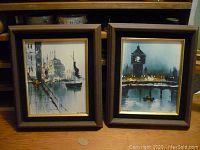 Pair of framed cityscape paintings on wooden surface