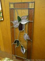 Full view of the rectangular stained glass panel with purple flower and brown border, hanging on a wooden wall.