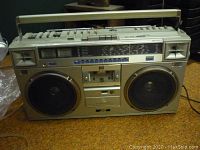 Front view of vintage JVC boom box showing dual large speakers, cassette player section, radio tuner display and control buttons and sliders on top.