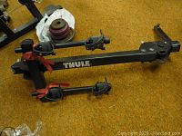 Full view of black metal Thule bike rack showing two arms with clamps and red protective straps, placed on carpeted floor.