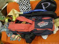 Photo showing a variety of sports equipment including a red and navy blue Molson hockey bag, yellow balls, a black and purple racket, pack of gloves with black spots, and some miscellaneous items on an orange surface.