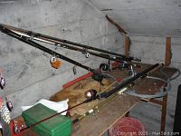 Wide view of six fishing rods arranged horizontally against concrete wall, showcasing different rod styles, handles, and reels attached.