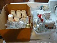 First image showing cardboard box filled with white teacups, clear glassware, and a brass bell, along with a tray holding more teacups with floral designs.