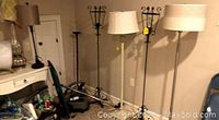Image showing three floor lamps with drum shades against a wall and two tall black metal floor candle holders beside them.