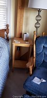 Small wooden accent table placed between a bed and a blue upholstered chair, showing its natural wood finish and open shelf space.