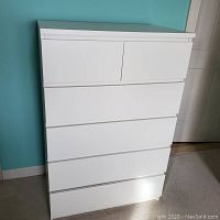Front view of the white tall dresser with five drawers showing smooth drawer fronts and the piece against a turquoise wall.