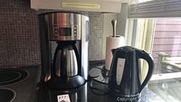 Black & Decker brushed metal coffee maker with glass carafe, next to black Sunbeam electric kettle on kitchen counter under window, with paper towel holder in background