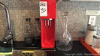 Red SodaStream soda maker unit on kitchen counter with two clear plastic SodaStream bottles behind it and clear glass decanter to the right.
