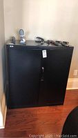 Front view of the black metal two-door lockable cabinet against a wall, with some items on top (not included).