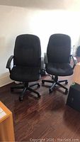 Front view of two matching black fabric office chairs with armrests and casters on wood floor.