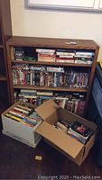 Full view of wooden book shelf filled with DVDs and VHS tapes on three shelves along with two cardboard boxes of additional media on floor