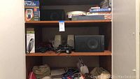 Shelf with electronic items including boxed hair clipper, two large black speakers, electronic accessories, and assorted boxed games.