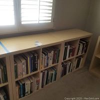 Two light wood finish Ikea Expedit style bookshelves each with 8 cubby compartments filled with assorted books under a window with shutters.
