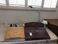 Two wood writing desks placed side by side on a surface, one light wood and one dark wood. Desk lamps and other objects partially visible in background.