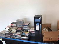 Assorted CDs and cassette tapes stacked on a flat table with Laser Line CD cabinet beside them.
