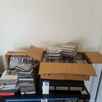 Wide view of two boxes and several stacks of CDs and cassette tapes on a table.