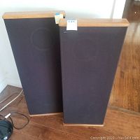 Two DCM speakers standing side by side on a wooden floor, front view with cloth grille covering.