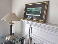 Ethan Allen composite and metal table lamp on glass table next to white mantel, with Douglas Busch signed framed landscape artwork resting on the mantel.