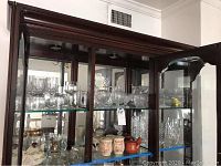 Full view of wooden display cabinet with multiple glass shelves holding various crystal and glassware items including stemmed glasses, lidded candy dishes, mugs, and bowls.
