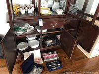 Wide angle view showing 5 Corning Ware casseroles stacked inside wooden cabinet, silver plate flatware chest open with serving ware inside, and additional flatware boxes on floor and shelf including boxed 100 Years collection