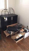 Ikea Expedit black shelving unit with wear, black desk lamp, black metal organizer racks, boxes on floor containing electronic parts and accessories including headphones, blank CDs, cables, printer option kit, battery backup and RCA receiver.