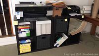 Two black Ikea Expedit shelving units with office supplies, printers, books, and boxes, showing wear.