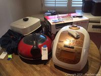 Photo of all four appliances together on table including HappyCall grill (red pan), Cuckoo rice cooker (white with digital display), Best Kitchen fish toaster box, and Presto cooker.