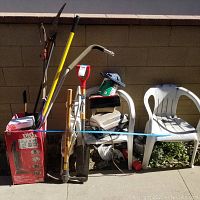Full view of assorted gardening tools and two white plastic chairs against a cinder block wall