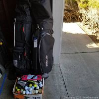 Image showing two black golf bags, one Yonex and one Real Feel, with clubs inside and a box of assorted golf balls and accessories in front.