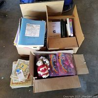 Box with assorted children’s books, spiral-bound notebooks, papers, small framed pieces, and plush toys including a white bear in heart sweater.