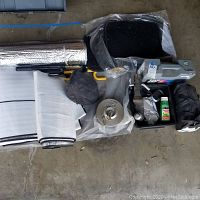 Wide shot showing assorted automotive supplies including tire chains in a torn black nylon bag, floor mats in packaging, a handheld tire chain tightening tool, emergency reflective triangle, brake cleaner, and various small items on concrete floor.