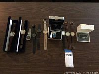 Wide shot of 9 various men's watches, some in display boxes, on wood table to show assortment and condition.