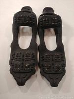 Pair of black rubber shoe ice grippers with multiple metal studs on bottom for ice traction, laid out on white tile floor.