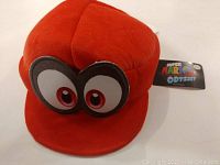 Red plush Super Mario Odyssey hat with large eyes front view and attached Super Mario Odyssey tag.