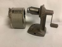 Gray metal rotary pencil sharpener labeled Adco Vacuhold with crank handle and suction base shown from angle view.