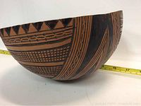 Side view of a hand carved and painted gourd bowl featuring intricate geometric black patterns on a natural brown background with a tape measure showing scale.