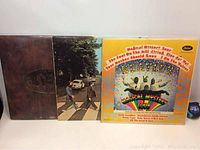 Three Beatles vinyl LP record albums standing side by side, showing front covers of Abbey Road, Magical Mystery Tour and the embossed brown cover album.