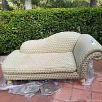 Front and side view of the beige chaise lounge with fleur-de-lis pattern upholstery and carved wood feet; note fabric texture and markings.