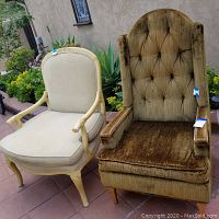 Two lounge chairs outside on patio: one cream painted wood with beige upholstery, one brown tufted velvet chair with wooden legs.